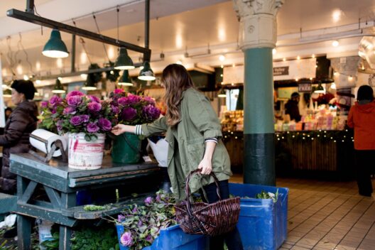 A woman with long hair and an olive green jacket selects flowers from a bucket at a bustling indoor market, holding a wicker basket and surrounded by purple blooms and shoppers.