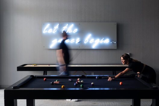 A woman prepares to take a shot at a pool table while a blurred man walks by. Behind them, a neon sign on the wall reads let the games begin.