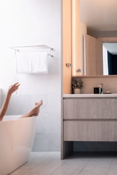 A person relaxing in a modern white bathtub, holding a drink. Only their legs and one arm are visible. The bathroom features light wood cabinets, white towels, and marble walls and floors.