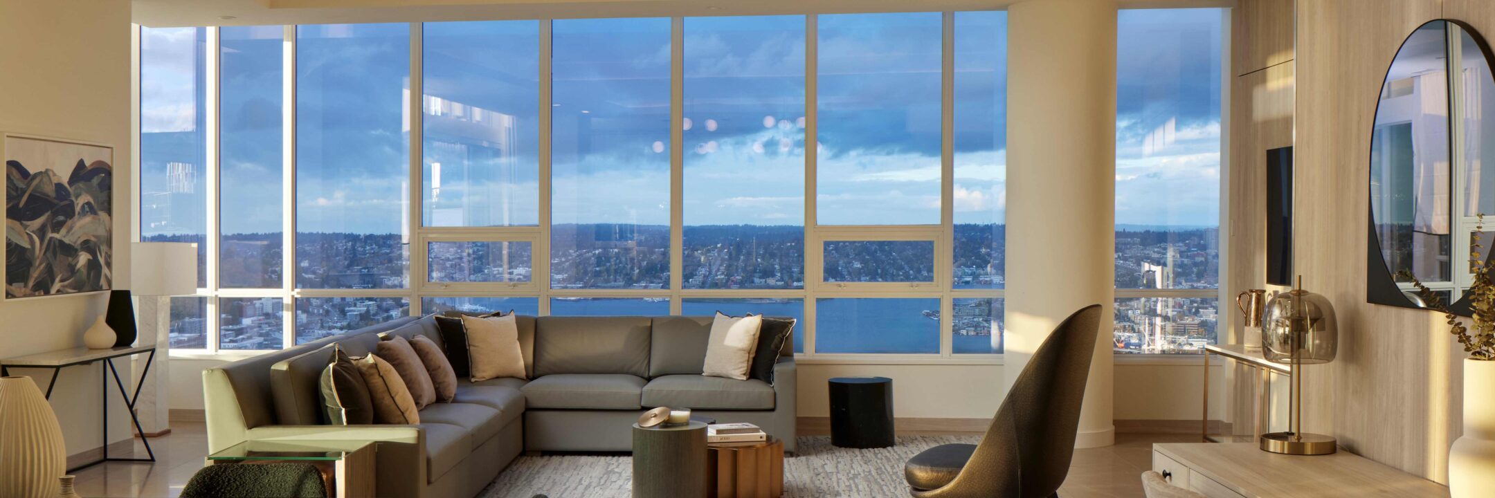 A modern living room with a large sectional sofa, chairs, and a coffee table, featuring floor-to-ceiling windows that offer a panoramic city and waterfront view under a blue sky.