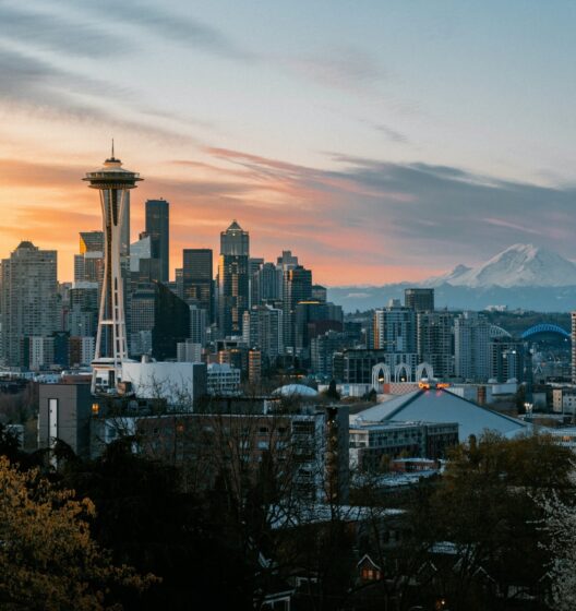 Seattle skyline at sunset with the Space Needle in the foreground, downtown skyscrapers, and Mount Rainier visible in the distance under a colorful sky.
