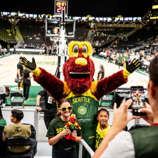 A person in a red and yellow mascot costume poses with excited kids in Seattle Storm shirts at a basketball arena, while someone takes their photo on a phone.