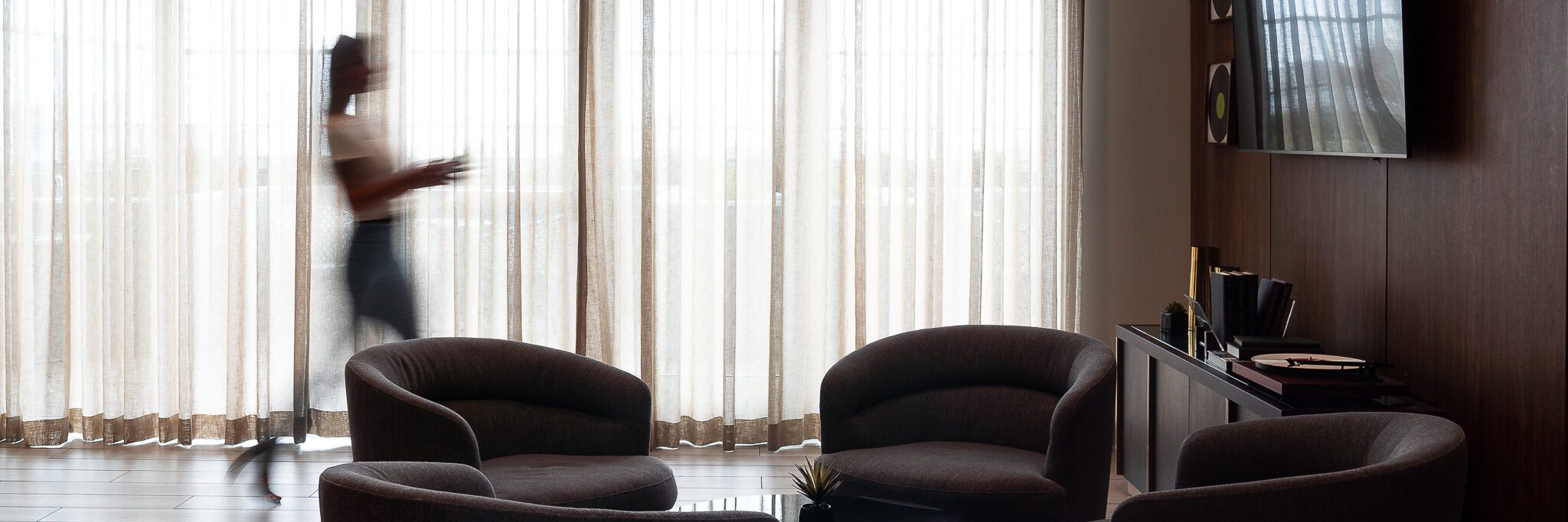 A blurred person walks past sheer floor-to-ceiling curtains in a modern lounge with three dark armchairs, a round table, and a sideboard with books and decor.