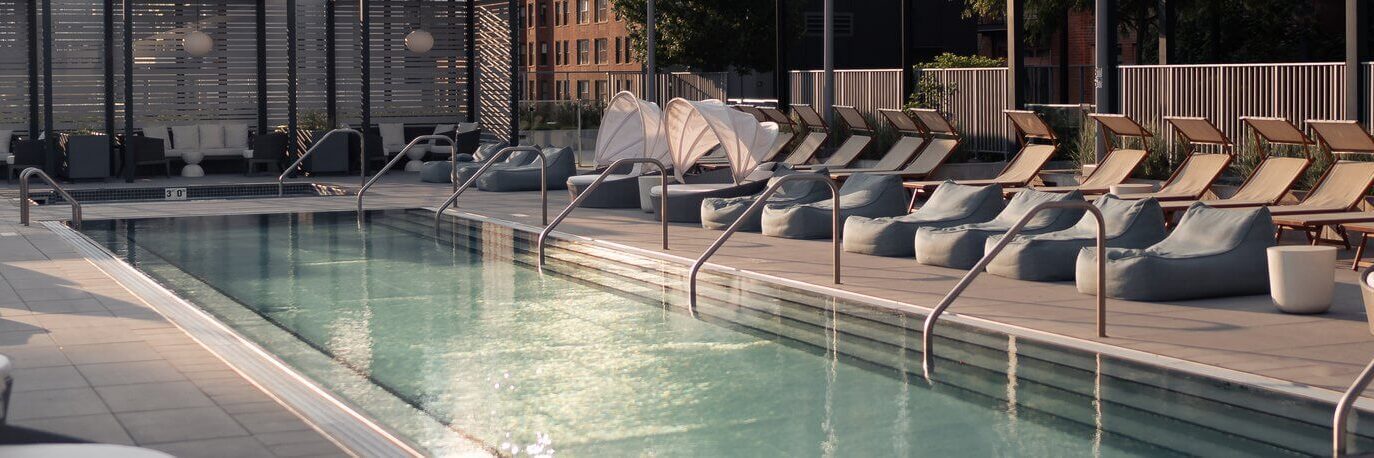 A modern outdoor pool surrounded by lounge chairs, bean bags, and shaded seating areas, with buildings and greenery in the background. The area appears calm and empty.