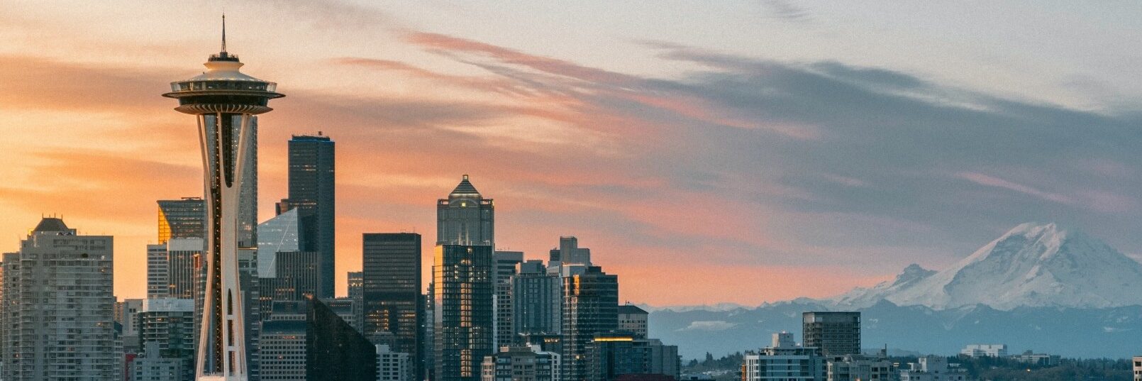 Seattle skyline at sunset with the Space Needle on the left, skyscrapers in the center, and Mount Rainier in the background under a sky with pastel clouds.