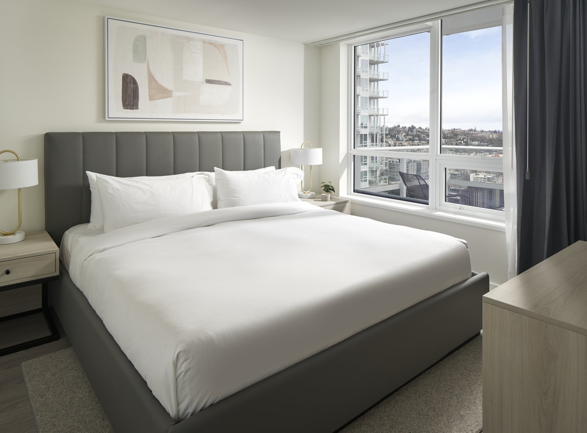 Modern bedroom with a large, neatly made bed with white bedding, two bedside tables with lamps, abstract art on the wall, large windows with city and sky views, and a dresser in the foreground.