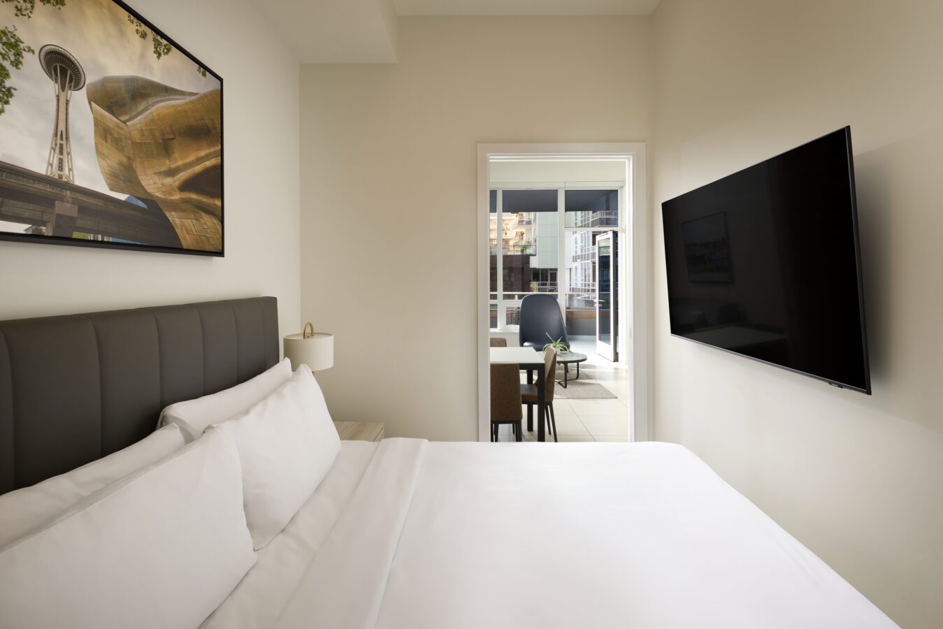 A modern hotel room with a neatly made bed, wall-mounted TV, artwork above the headboard, and a view into a workspace with a desk and chair by a window.