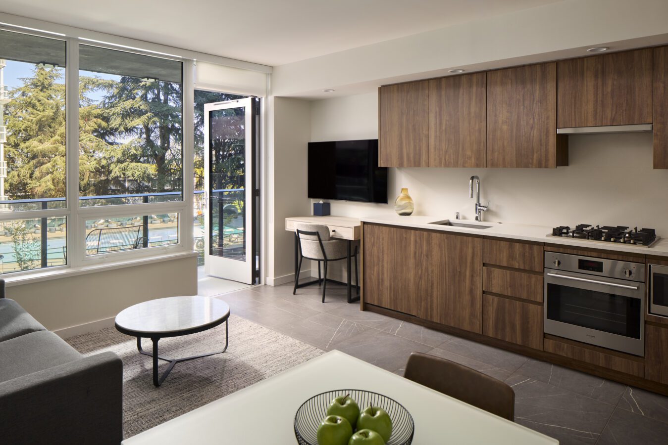 Modern apartment with wood cabinets, a kitchen, desk, TV, sofa, round table, and bowl of green apples. Large windows and open door show greenery and sunlight outside. Neutral tones dominate the décor.