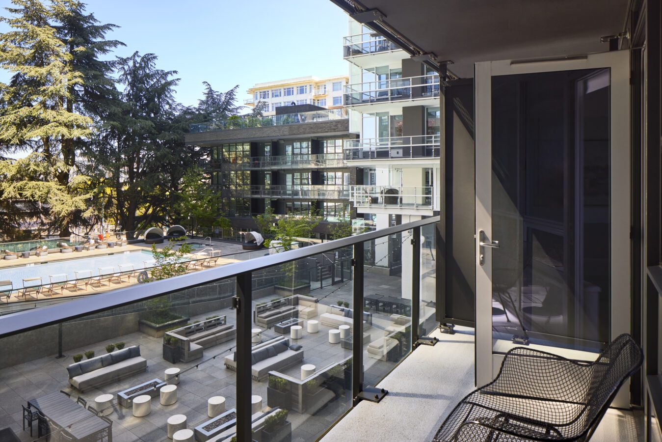 A modern apartment balcony with two chairs overlooks a courtyard with seating areas, plants, and a pool. Glass railings line the balcony, and other apartment buildings are visible in the background.