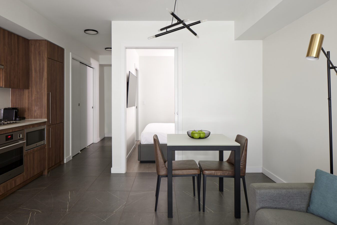 Modern apartment interior with a small dining table and two chairs, a kitchen with wood cabinets on the left, a minimalist light fixture, and a bedroom visible through an open doorway in the background.