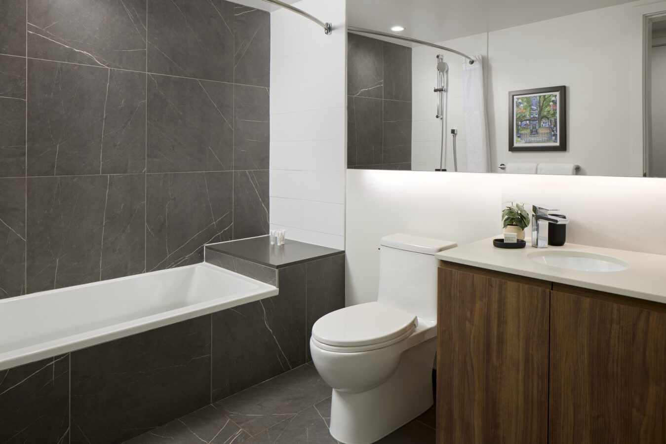 Modern bathroom with a bathtub, white toilet, wood vanity, large mirror, small plant, and gray marble tile walls. A framed picture hangs on the white wall above the bathtub.
