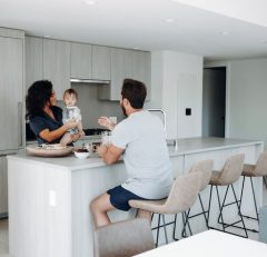 A woman holds a baby while talking to a man seated at a modern kitchen island. The kitchen features light wood cabinets, bar stools, and minimalistic decor.