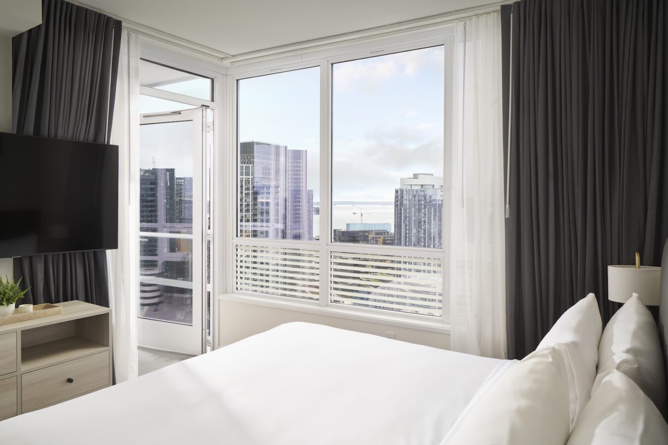 A modern hotel room with a neatly made bed, a wall-mounted TV, and a large window showing a city skyline with tall buildings. The balcony door is open, allowing natural light into the bright, clean space.