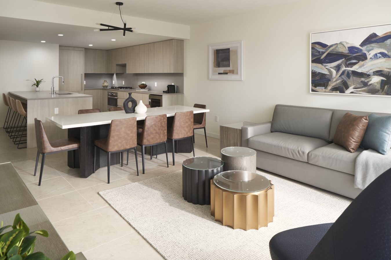 Modern open-concept kitchen and living room with light wood cabinets, a long island with brown barstools, a dining table with leather chairs, a gray sofa, abstract art, and stylish metallic coffee tables on a rug.