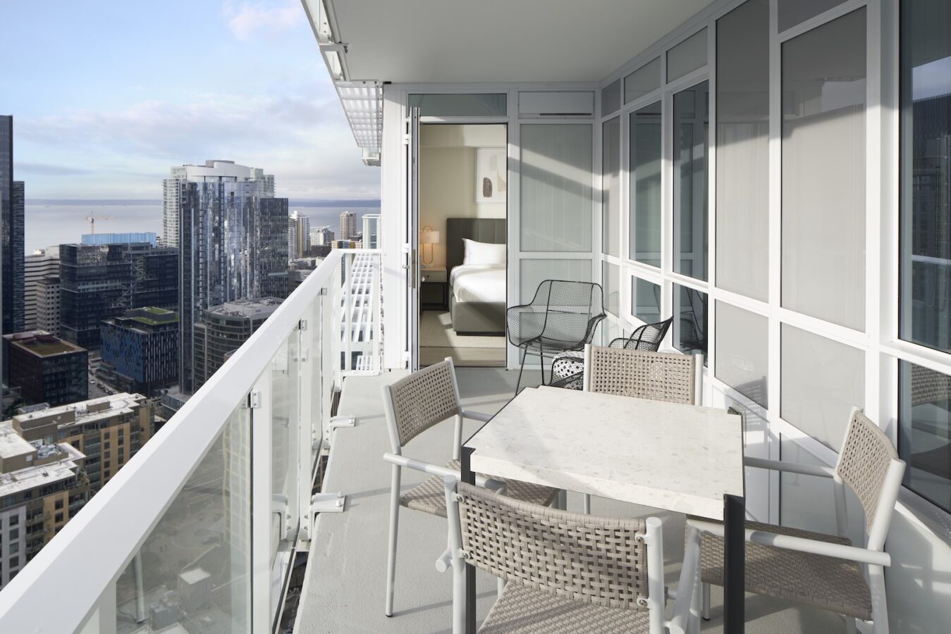 A modern high-rise balcony with a table and chairs overlooks a city skyline with tall buildings and distant water. The balcony connects to a sleek, glass-walled bedroom with a neatly made bed and minimal decor.