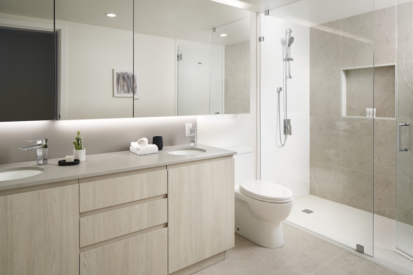 Modern bathroom with light wood cabinetry, double sinks, a large mirror, a white toilet, and a spacious glass-enclosed shower with beige tiles. Minimalist decor includes a small plant and folded towels on the counter.