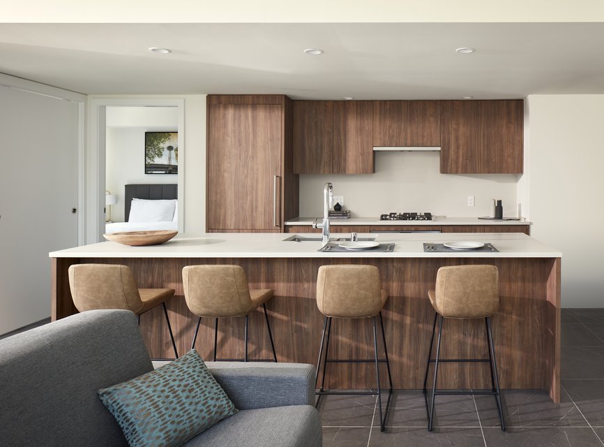 Modern kitchen with wood cabinets and island, four tan barstools, built-in stove, and sink. Bedroom visible through open door; gray sofa with patterned pillow in foreground. Bright, clean, and minimalist design.