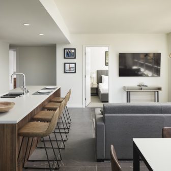 Modern, open-concept apartment with a kitchen island and barstools, a gray sofa facing a wall-mounted TV, a dining table, and a bedroom visible through an open doorway. Decor is minimalist with neutral colors.
