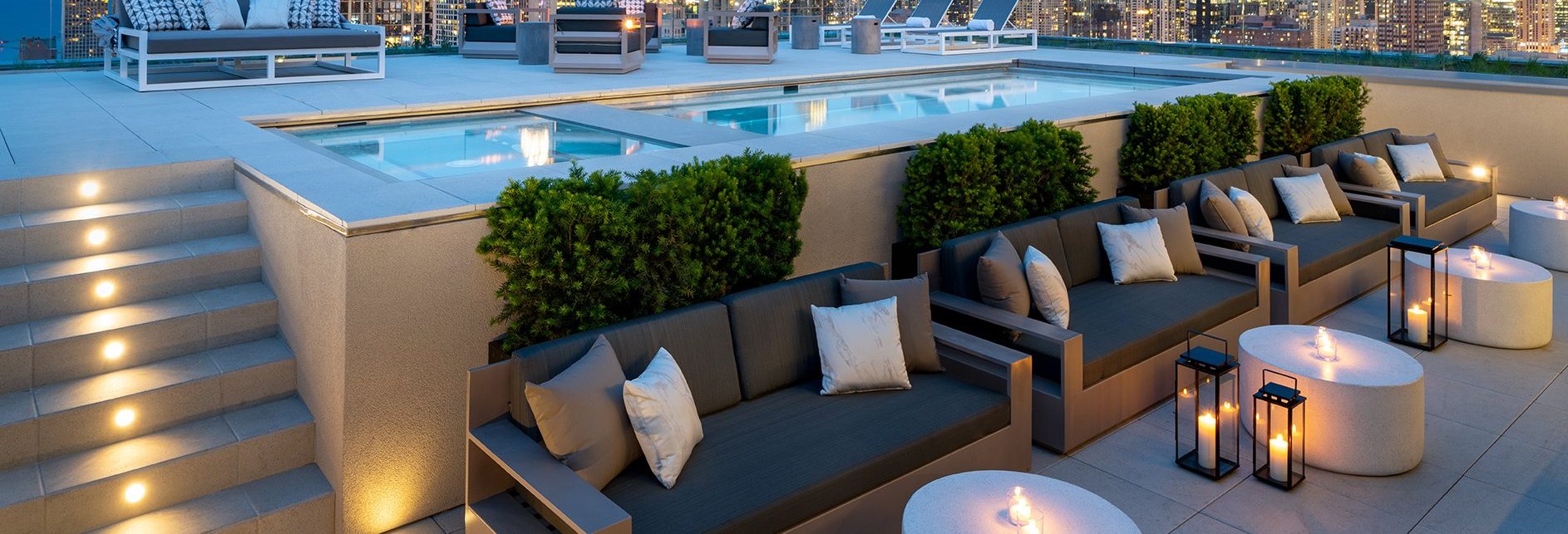 A rooftop lounge area at night features modern sofas with cushions, round tables with lit candles, lanterns, a swimming pool, and views of a city skyline in the background. Steps with lights lead up to the pool area.