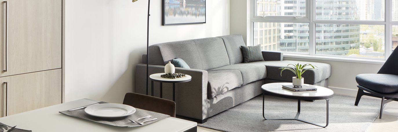 Modern living room with a gray sectional sofa, two round coffee tables, a potted plant, and large windows showing city buildings; a dining table with a place setting is in the foreground.