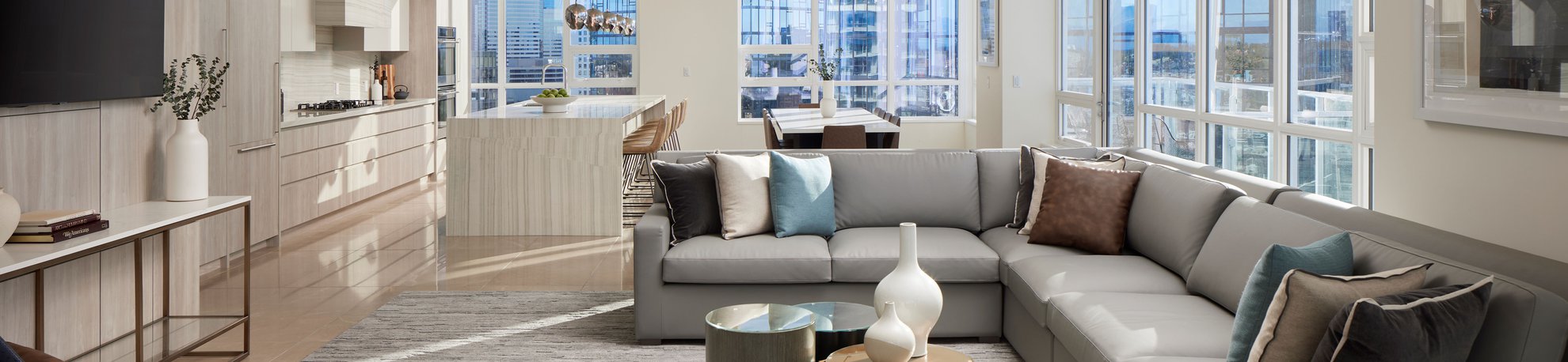 Modern, sunlit open-plan living area with a large gray sectional sofa, glass coffee tables, and a kitchen with bar stools. Floor-to-ceiling windows offer city views, filling the space with natural light.