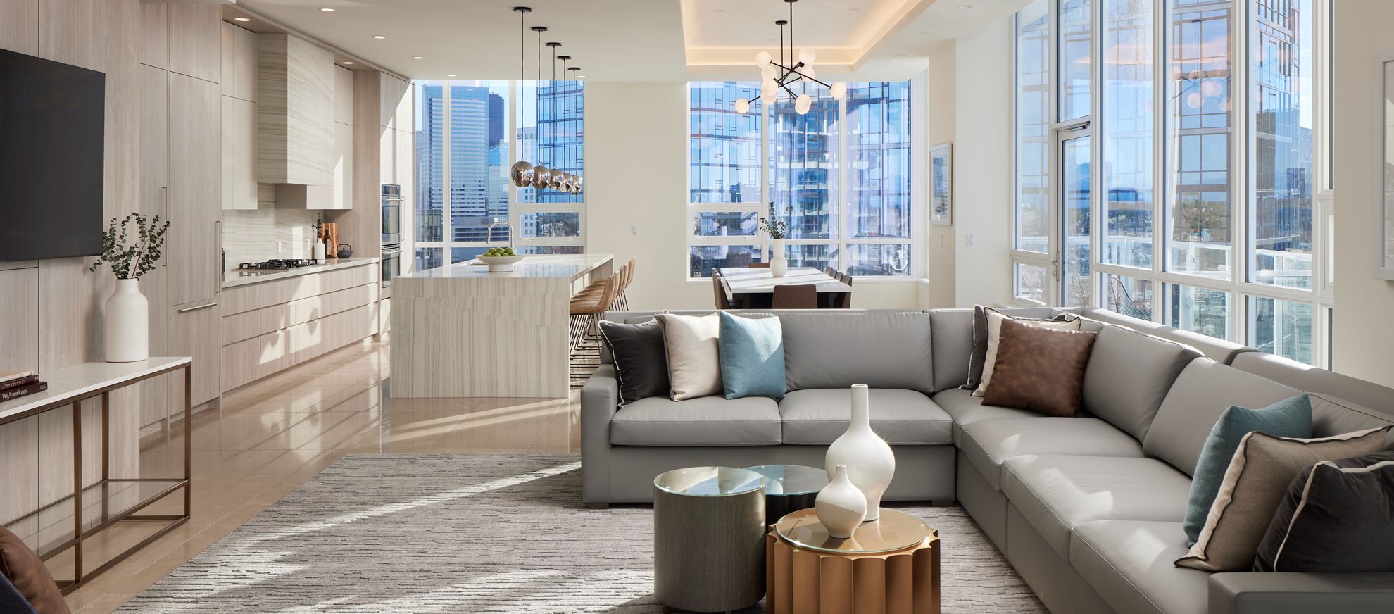 Modern open-concept living space with a gray sectional sofa, stylish decor, a kitchen with an island, dining area, and large floor-to-ceiling windows offering city views and ample natural light.