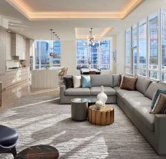 A modern, sunlit living room with a gray sectional sofa, stylish tables, decorative vases, large windows with city views, and an open kitchen in the background. Neutral tones and contemporary decor create an inviting space.