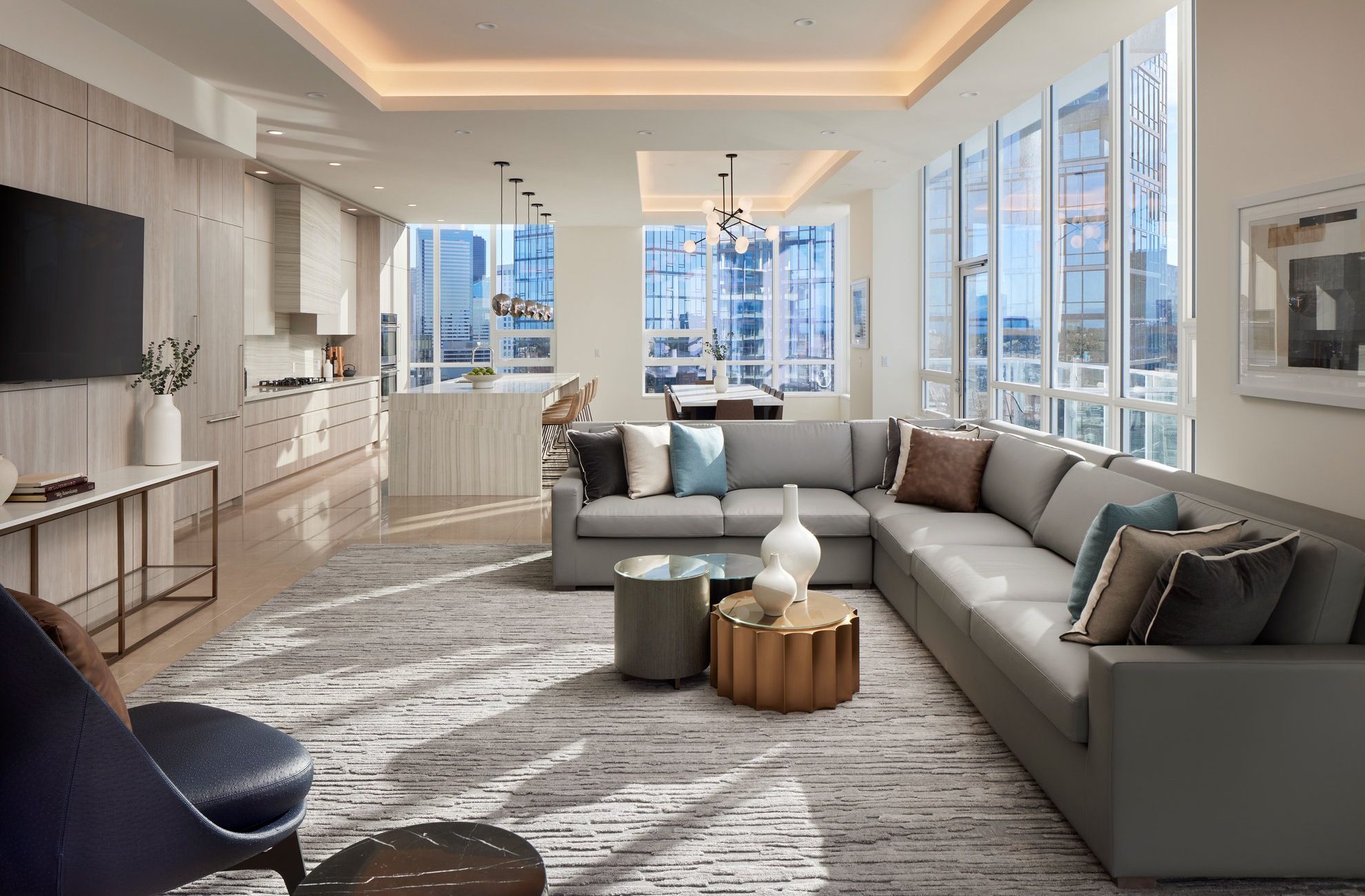 A modern, sunlit living room with a large gray sectional sofa, stylish armchairs, a gold accent table, and floor-to-ceiling windows offering city views. Light wood cabinets line the open kitchen in the background.