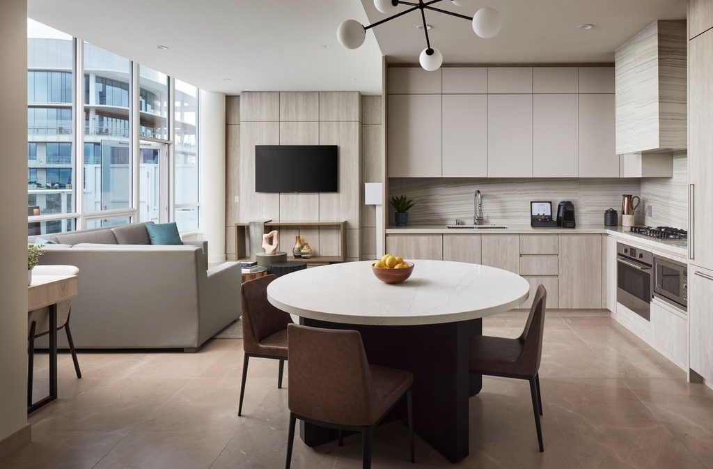 Modern open-concept kitchen and living area with neutral tones, round dining table with four chairs, built-in appliances, wall-mounted TV, and large windows providing natural light and city views.