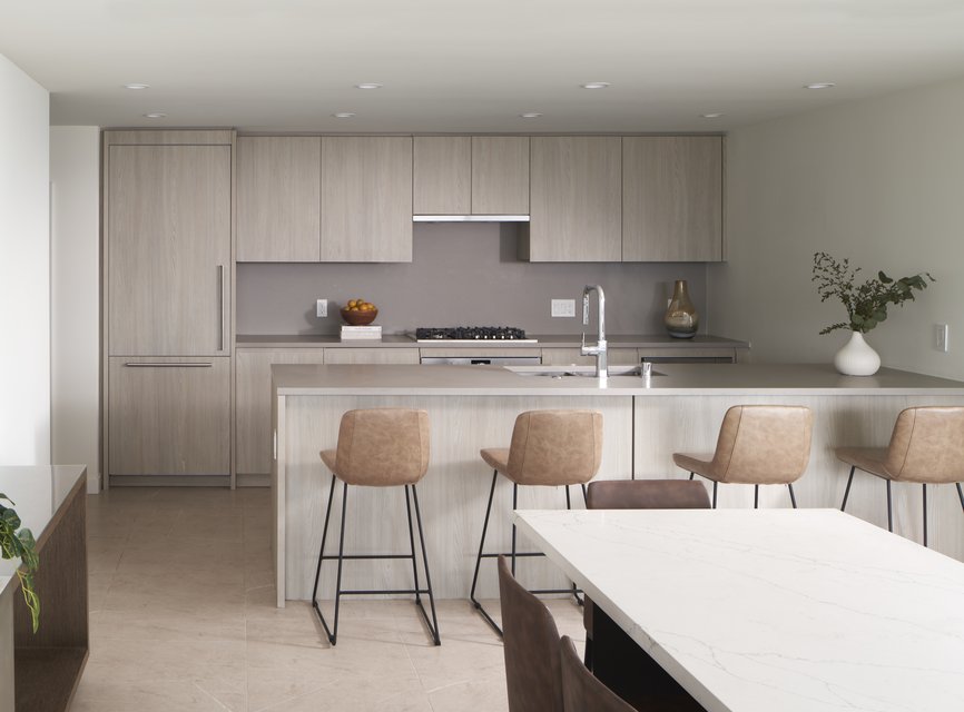 Modern kitchen with light wood cabinets, integrated appliances, and a central island with four tan barstools. A white countertop table and minimalist decor complete the bright, clean space.