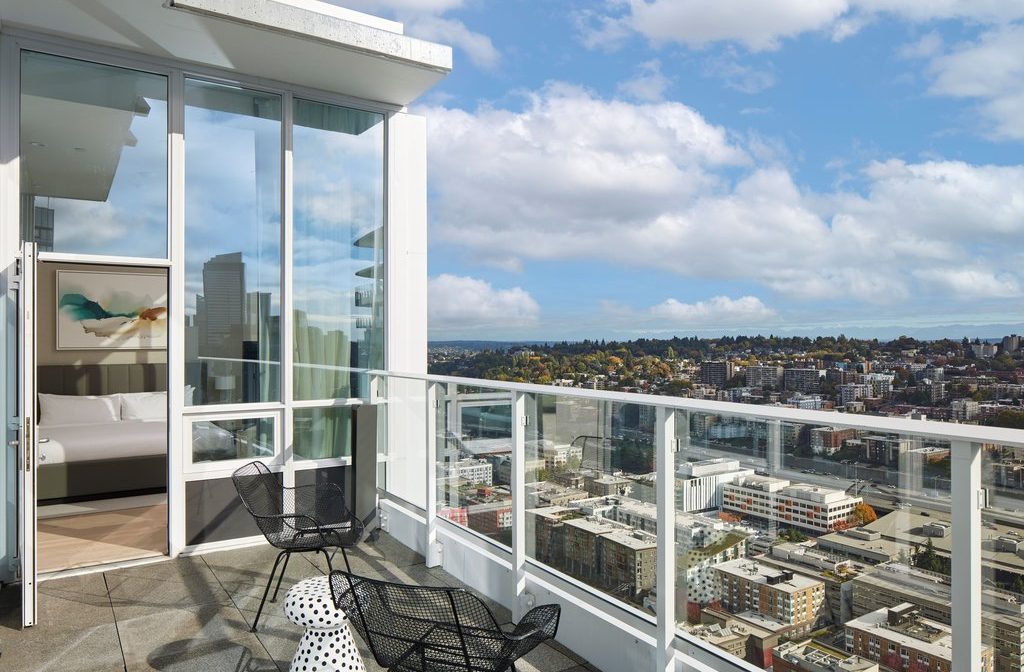 A modern balcony with glass railings features black chairs and a small table, overlooking a cityscape of buildings, trees, and distant hills under a partly cloudy sky. An open door leads to a bright bedroom inside.