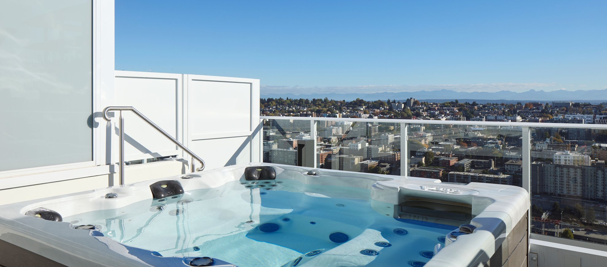A rooftop hot tub with clear water overlooks a cityscape under a bright blue sky. Glass railings provide a view of buildings, trees, and distant mountains.