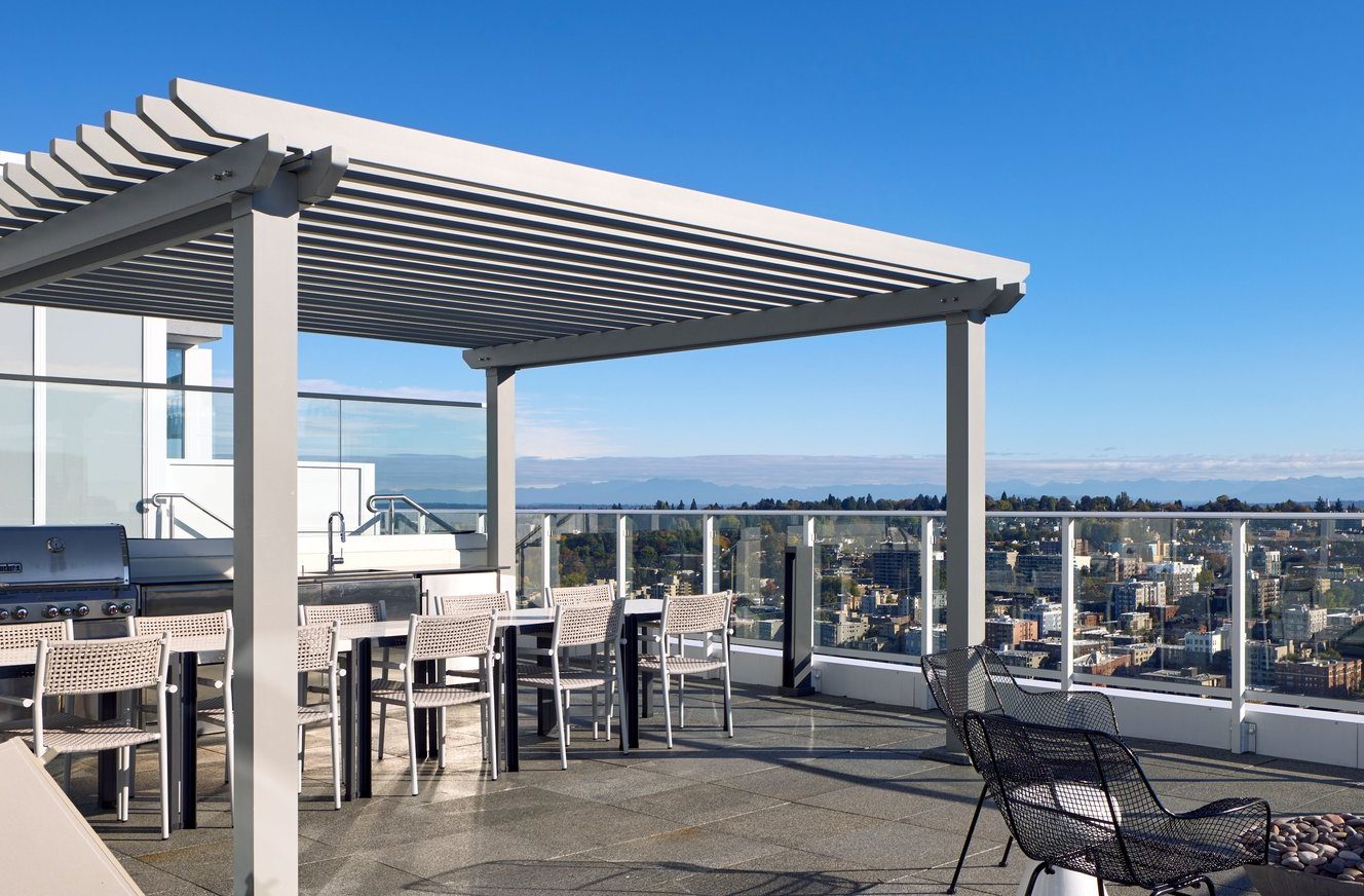A rooftop patio with a pergola covering a long dining table and chairs, a barbecue grill, lounge chairs, and glass railings overlooking a cityscape and distant mountains under a clear blue sky.