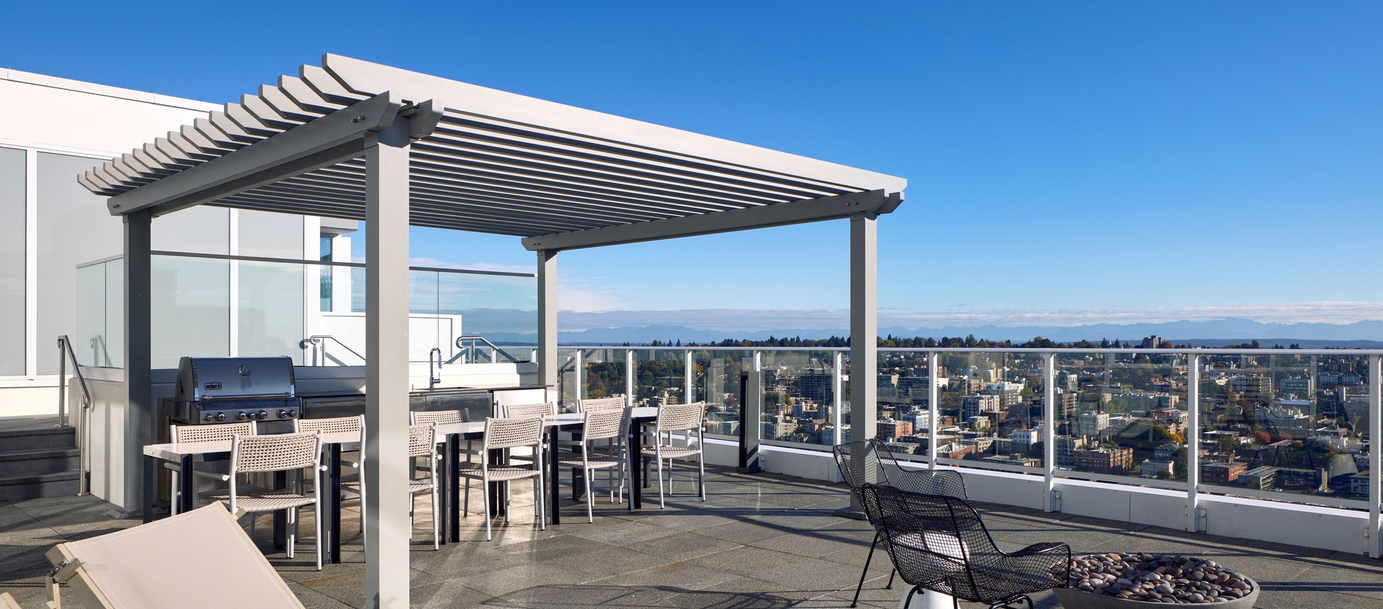 A modern rooftop patio with a pergola, a dining table and chairs, a grill, lounge chairs, and glass railings offers a city view under a clear blue sky.