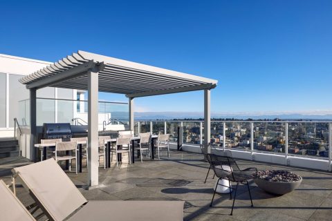 A modern rooftop patio with lounge chairs, a pergola over a dining area, a barbecue grill, black lounge chairs, a fire pit, and glass railings overlooking a cityscape under a clear blue sky.