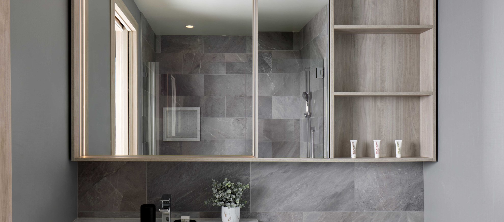 Modern bathroom with gray stone tiles, a large mirrored cabinet, open wooden shelves with toiletries, a glass shower door, and a countertop with a small potted plant and various items.