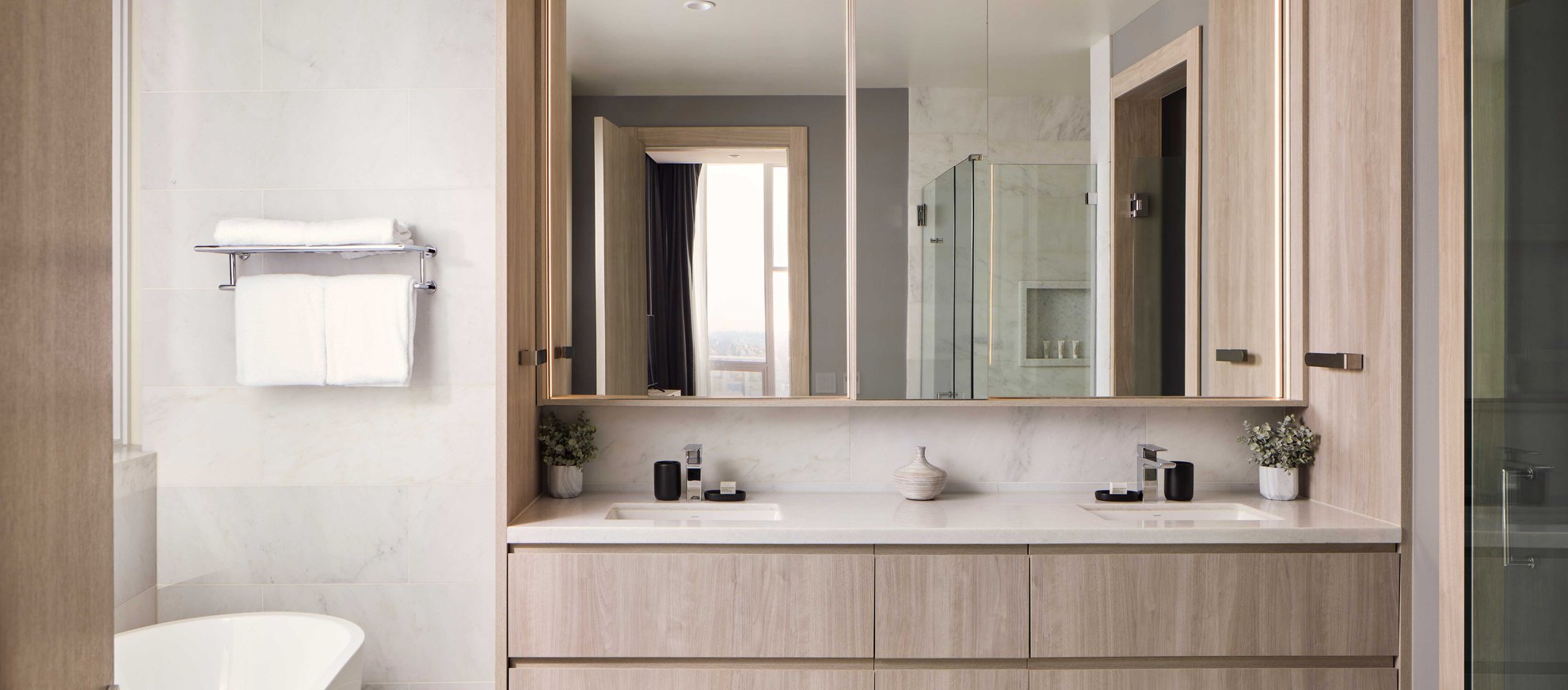 Modern bathroom with light wood cabinets, a double sink vanity, large mirrors, a white towel on a rack, a bathtub, and glass shower. Minimalist decor with small plants and toiletries on the counter.