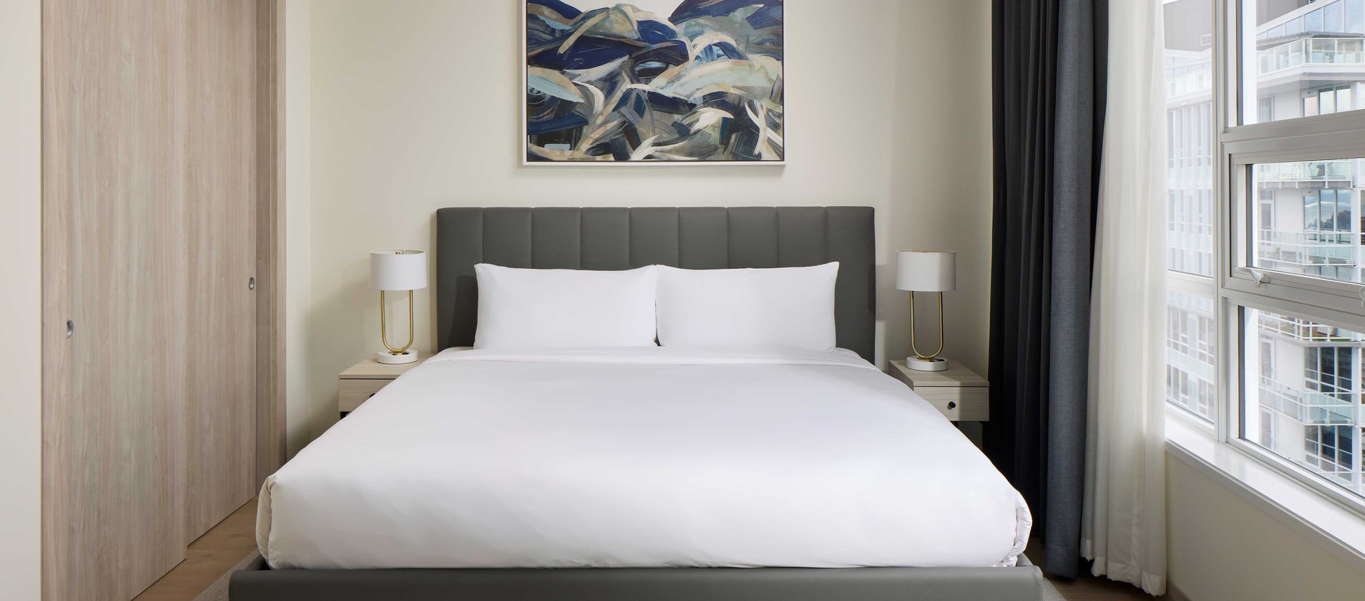 A modern bedroom featuring a neatly made bed with white linens, two bedside tables with lamps, a large abstract painting above the bed, and a window with city views and dark curtains.