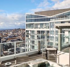 A rooftop terrace with seating, a hot tub, and glass railings overlooks a cityscape with modern buildings. The Space Needle and water are visible in the distance under a partly cloudy sky.