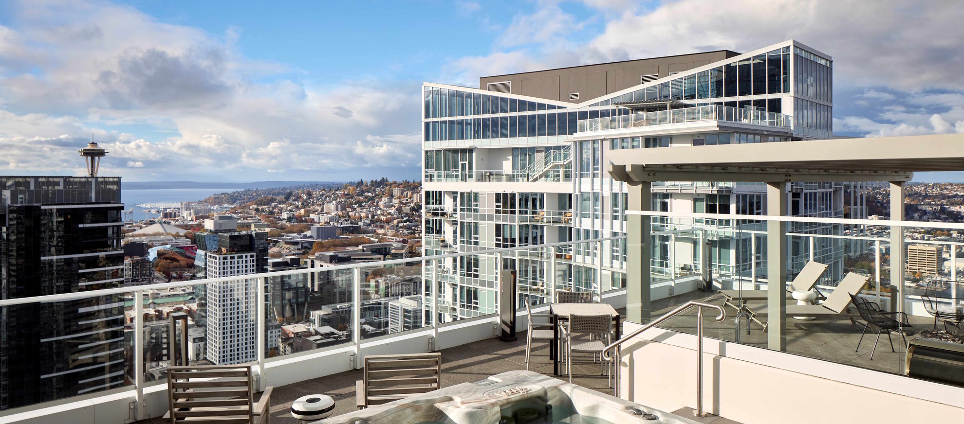 A rooftop terrace with seating, a hot tub, and glass railings overlooks a cityscape with modern buildings. The Space Needle and water are visible in the distance under a partly cloudy sky.