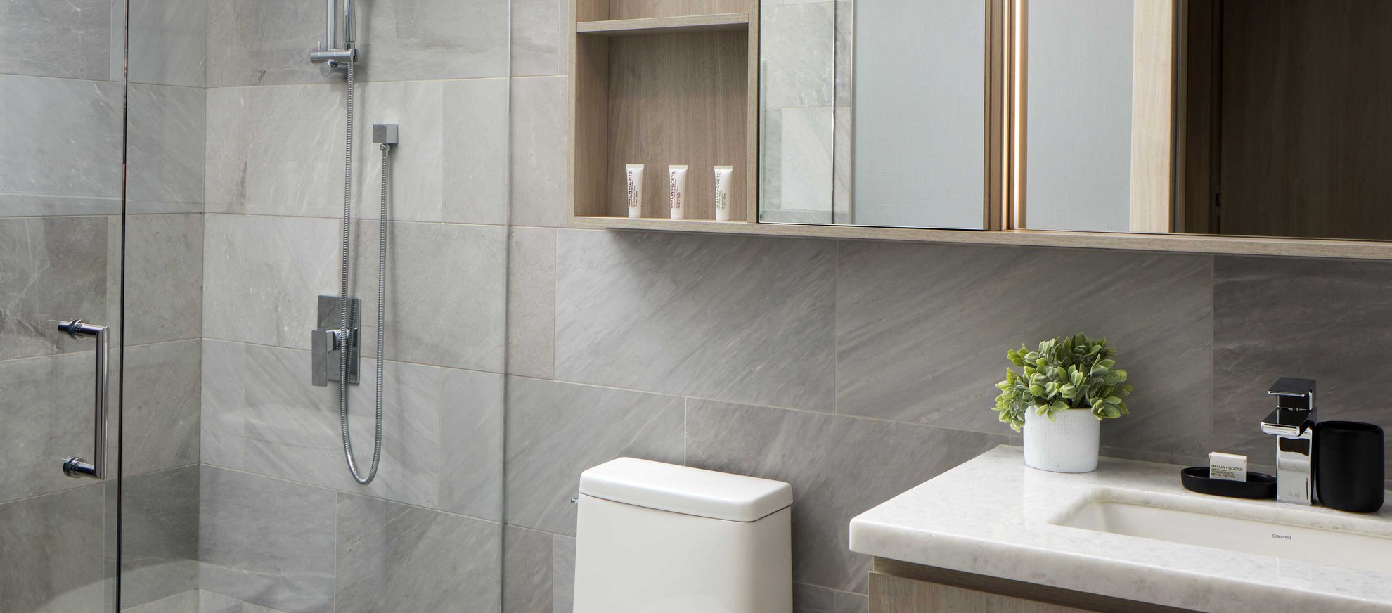 Modern bathroom with gray tiled walls, a glass shower door, a white toilet, a sink with a countertop, a potted plant, toiletries, and mirrored cabinets above the sink.