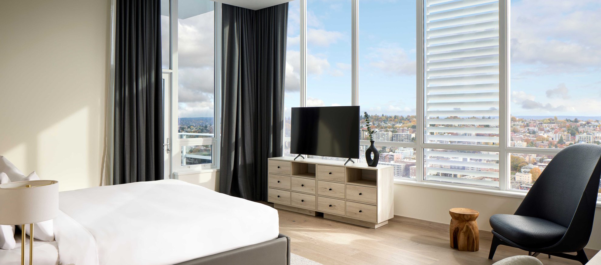 A modern bedroom with a large bed, a dresser with a TV, a chair, a small round wooden table, and floor-to-ceiling windows offering a city view under a partly cloudy sky. Dark curtains frame the windows.