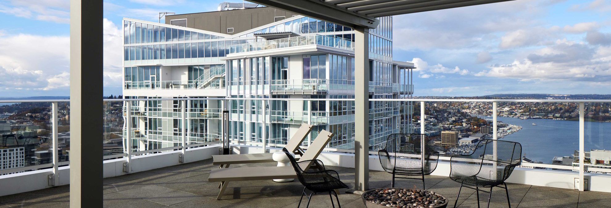A modern rooftop patio with lounge chairs, wire chairs, and a fire pit overlooks a city skyline, glass buildings, and a large body of water under a partly cloudy sky.
