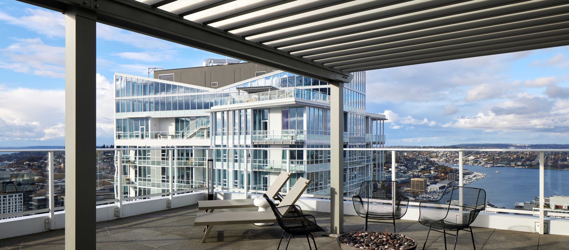 A rooftop patio with modern outdoor seating, a fire pit, and a pergola overlooks a cityscape with glass buildings, a body of water, and partly cloudy skies in the background.
