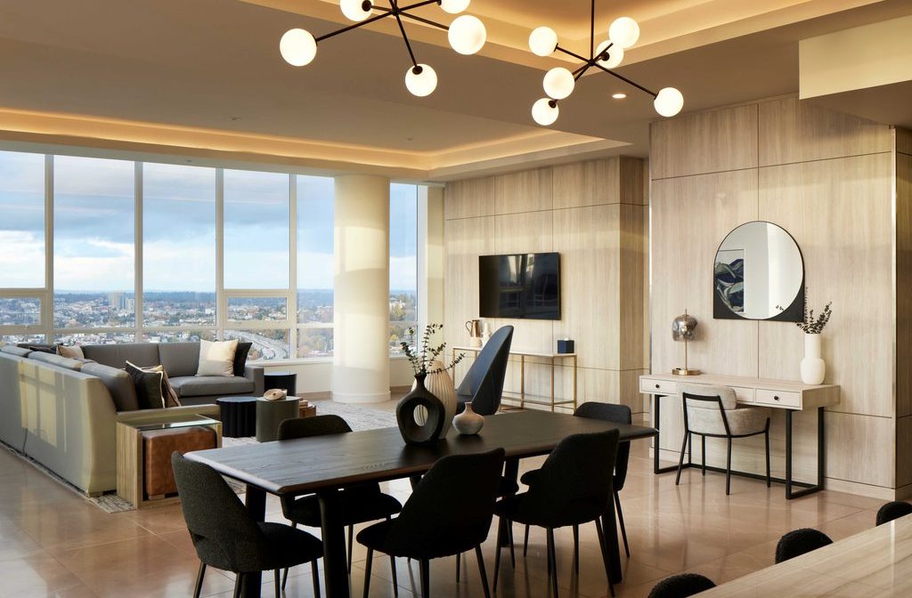 Modern open-concept living and dining area with large windows, city view, neutral tones, contemporary furniture, a wall-mounted TV, round mirror, and decorative lighting fixture overhead.