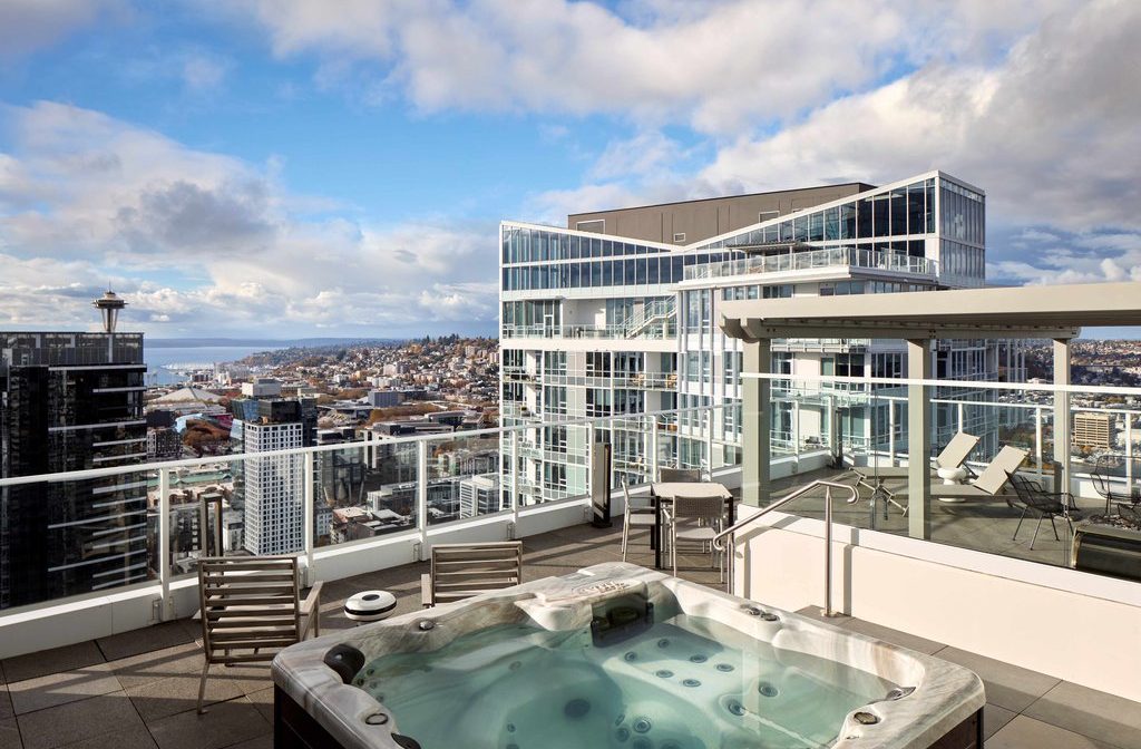 A rooftop patio with a hot tub, lounge chairs, and glass railings overlooks a cityscape with modern high-rise buildings, distant hills, and partly cloudy skies. The Space Needle is visible in the background.