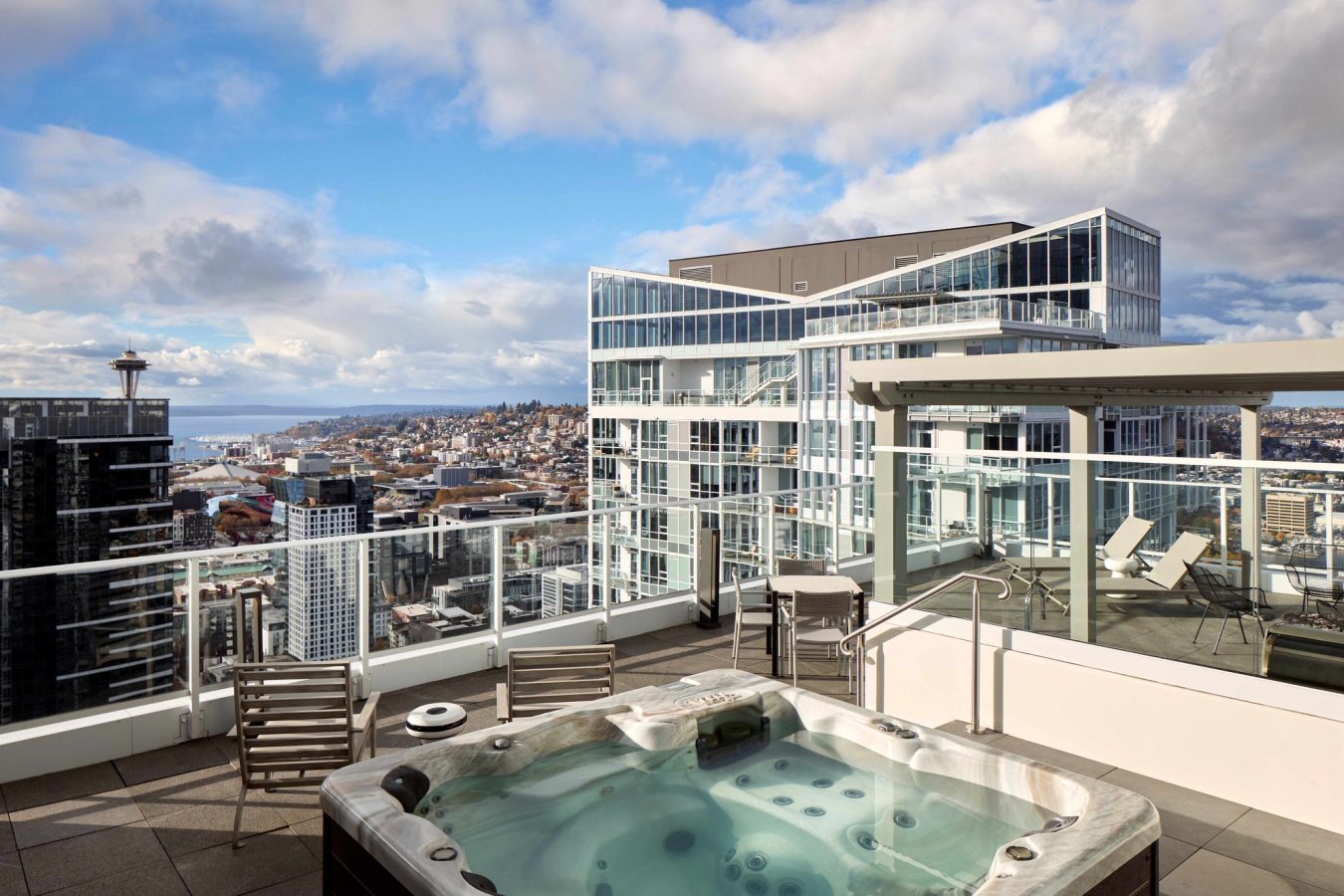 A rooftop patio features a hot tub and seating area, surrounded by glass railings. Modern high-rise buildings and a distant view of the Space Needle and cityscape are visible under a partly cloudy sky.
