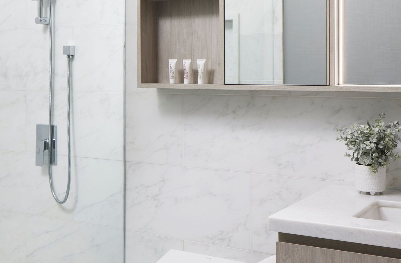 Modern bathroom with white marble walls, a built-in shower featuring a handheld showerhead, a wooden cabinet with toiletries, a white sink with a light countertop, and a small potted plant.