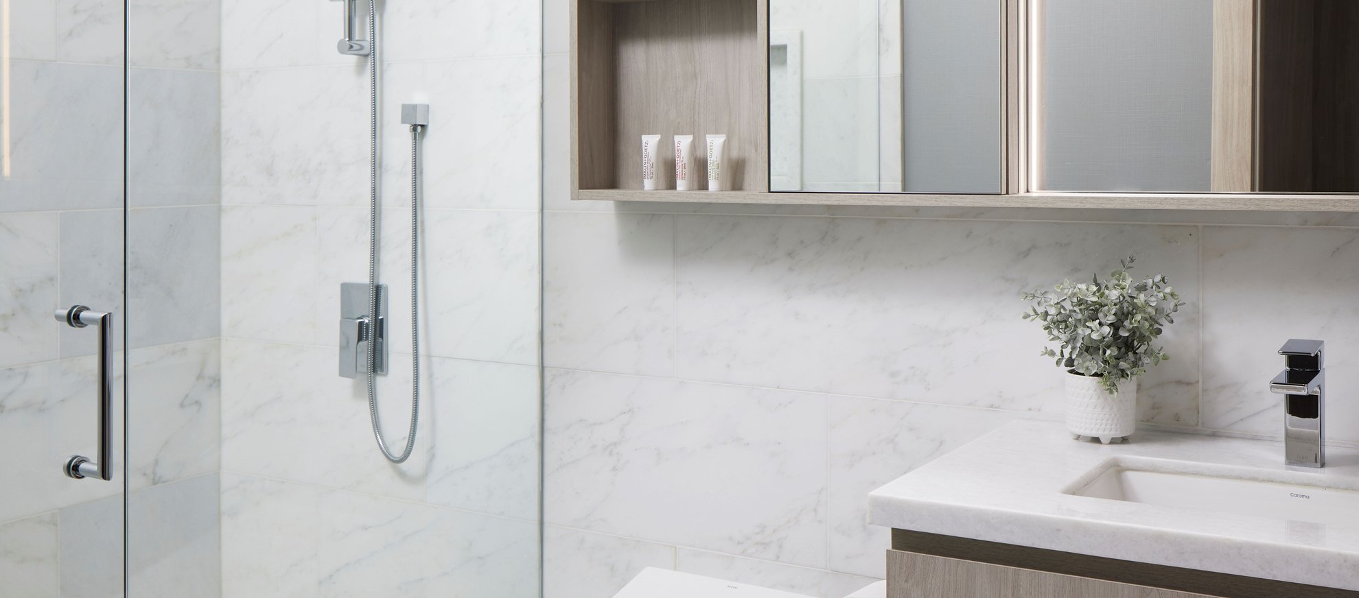 Modern bathroom with a glass shower, marble walls, mounted shower head, vanity with sink, faucet, mirror cabinets, small plant, and toiletries on a shelf. Clean, minimal design in neutral tones.