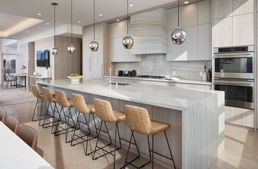 Modern kitchen with a large marble island, five tan leather barstools, pendant lights, built-in oven, light wood cabinets, gas cooktop, and a bowl of green apples on the counter. Dining and living areas are visible in the background.